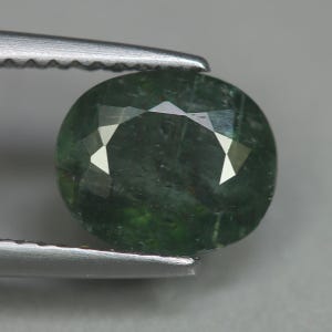 May include: A faceted, oval-shaped gemstone with a deep green hue. The gem is held by a pair of silver tweezers, showcasing its polished surfaces and internal clarity. The stone's facets reflect light, creating bright highlights.