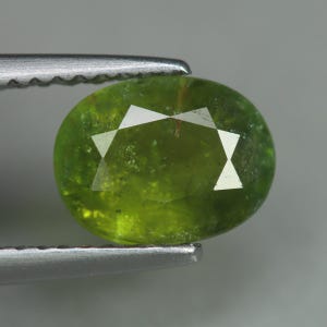 May include: A faceted, oval-shaped, green gemstone held by a pair of silver tweezers. The gem has a vibrant, translucent appearance with a star-like pattern of facets on its surface. The background is a neutral gray.