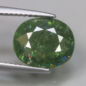May include: A faceted, oval-cut green gemstone held by silver tweezers. The gem has a vibrant, translucent green color with internal inclusions and iridescent flashes. The cut features a star-like pattern.