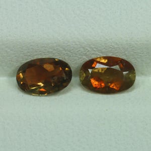 May include: Two oval-cut gemstones with a warm, amber color. The gems have a faceted surface that catches the light, displaying a mix of orange, brown, and yellow hues. The stones are set against a neutral background.