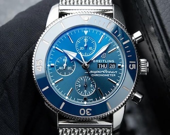 Könnte beinhalten: Eine Breitling Superocean Chronometer Uhr mit blauem Sonnenschliff-Zifferblatt, drei Hilfszifferblättern und einer Tages-Datums-Anzeige auf der Drei-Uhr-Position. Die Uhr verfügt über ein silberfarbenes Edelstahl-Mesh-Armband und eine drehbare blaue Keramiklünette.