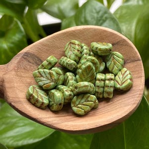 May include: A wooden spoon holds a collection of green leaf-shaped beads with brown veining. The beads are a mix of sizes and shapes, with a glossy finish. The background features lush green foliage.
