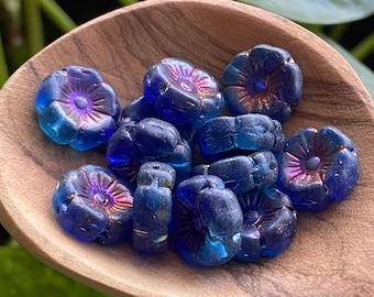 May include: A collection of dark blue flower-shaped beads with iridescent accents, displayed in a wooden scoop. The beads have a detailed floral design with a purple center and are suitable for jewelry making or craft projects.