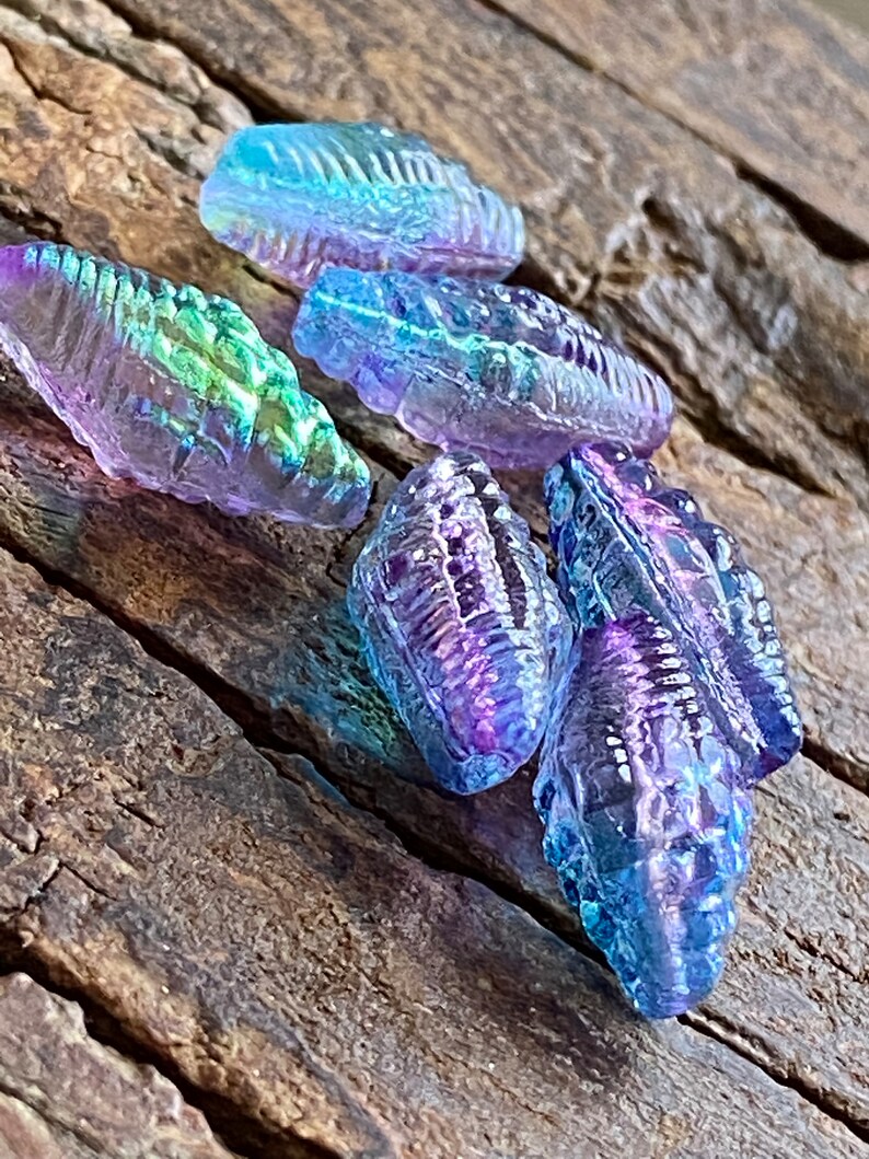 May include: Five iridescent glass beads in shades of purple, blue, and green. The beads are shaped like seashells and have a textured surface.