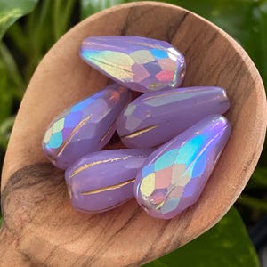 May include: Five teardrop-shaped lavender beads with a faceted surface and an iridescent finish. Each bead features a distinct gold-colored line running vertically along the side, creating a textured appearance on the light purple glass surface.