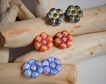 May include: A collection of flower-shaped stud earrings in various colors. The earrings are in pairs, with colors including blue with gold accents, orange with pink highlights, and iridescent purple. The earrings are displayed on driftwood.