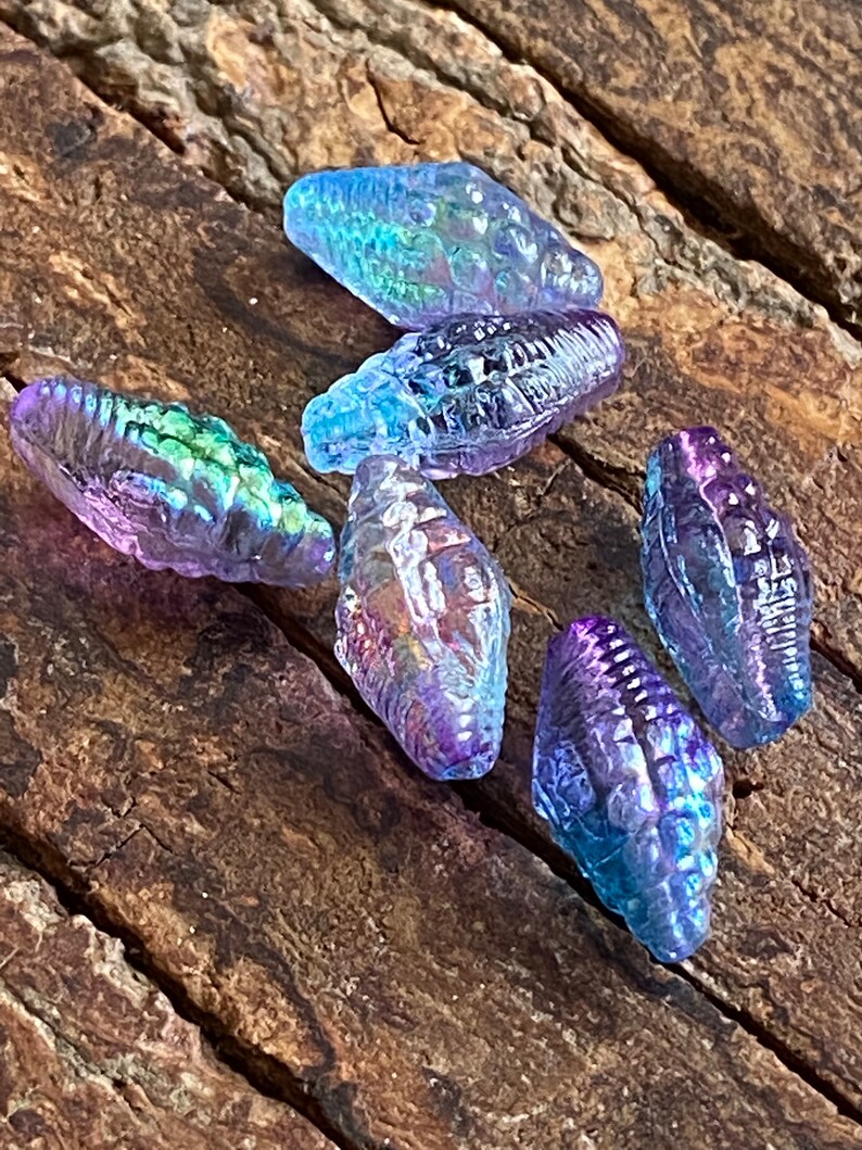 May include: Six iridescent glass beads in the shape of seashells. The beads are a mix of purple, blue, and green.