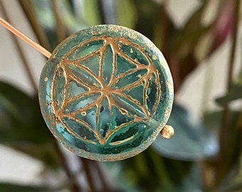 May include: A round, teal-colored bead with a gold-colored, etched floral design. The bead has a slightly textured surface and is strung on a thin, copper-colored wire. The background is blurred, showing green foliage.