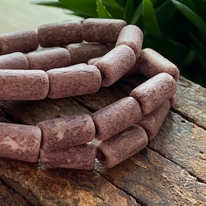May include: A close-up of a stack of mauve-colored ceramic beads with a rough, textured surface. The beads are cylindrical in shape and have a slightly irregular form.