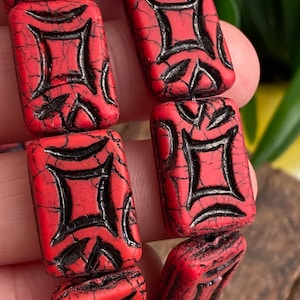 May include: Close-up of a red bracelet with rectangular beads. Each bead features a black geometric design and a cracked, textured surface. The bracelet is held in a hand, showcasing the details of the jewelry.