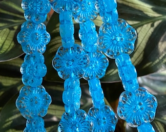 Könnte beinhalten: Nahaufnahme von Strängen aus hellblauen Glasperlen. Jede Perle hat die Form einer Blume mit detaillierten Blütenblättern und einem zentralen Design. Die Perlen sind aufgefädelt und bilden ein zartes und kompliziertes Muster. Der Hintergrund ist verschwommen grün.
