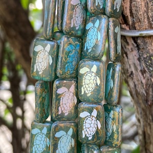 May include: A strand of rectangular beads in shades of green and gold. Each bead features a white sea turtle design. The beads are strung together, creating a decorative element. The background is blurred, suggesting an outdoor setting.