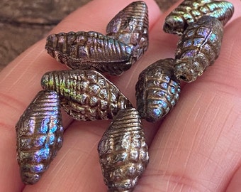 May include: Close-up of several small, metallic-looking, cone-shaped beads. The beads have a textured surface and iridescent color variations of blue, purple, and green. The beads are held in a person's hand.