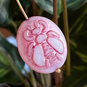 May include: A pink glass bead with a raised, detailed design of a stylized insect. The bead has a white center and a pink, iridescent finish.