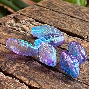 May include: Five iridescent glass beads in shades of purple and blue. The beads are shaped like teardrops and have a textured surface.