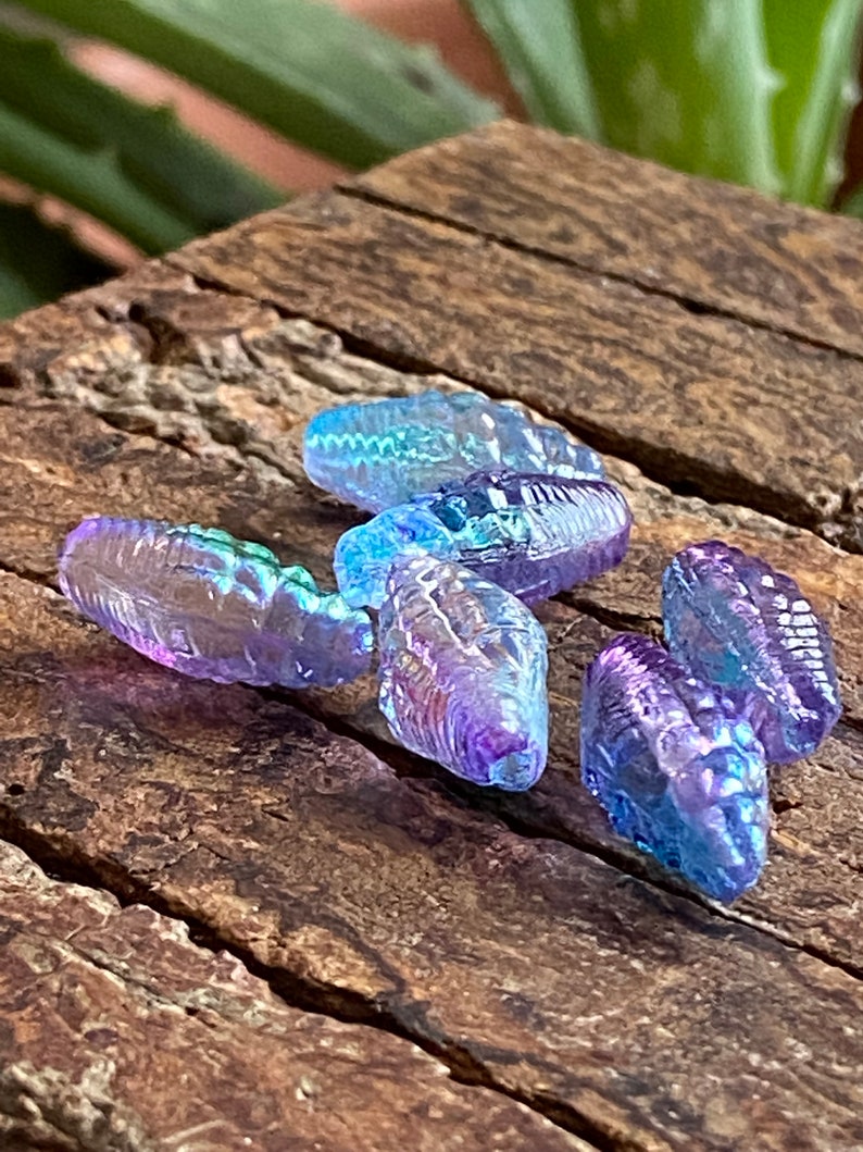 May include: Five iridescent glass beads in shades of purple and blue. The beads are shaped like teardrops and have a textured surface.