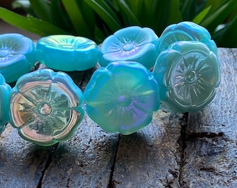 May include: Close-up of several iridescent blue flower-shaped beads. The beads have a glossy finish and are arranged on a weathered wooden surface. The flower beads have a detailed petal design and a central floral pattern.