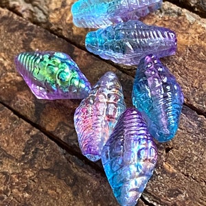 May include: A collection of iridescent glass seashell beads in shades of blue, purple, and green. The beads are all different shapes and sizes, and they have a textured surface.