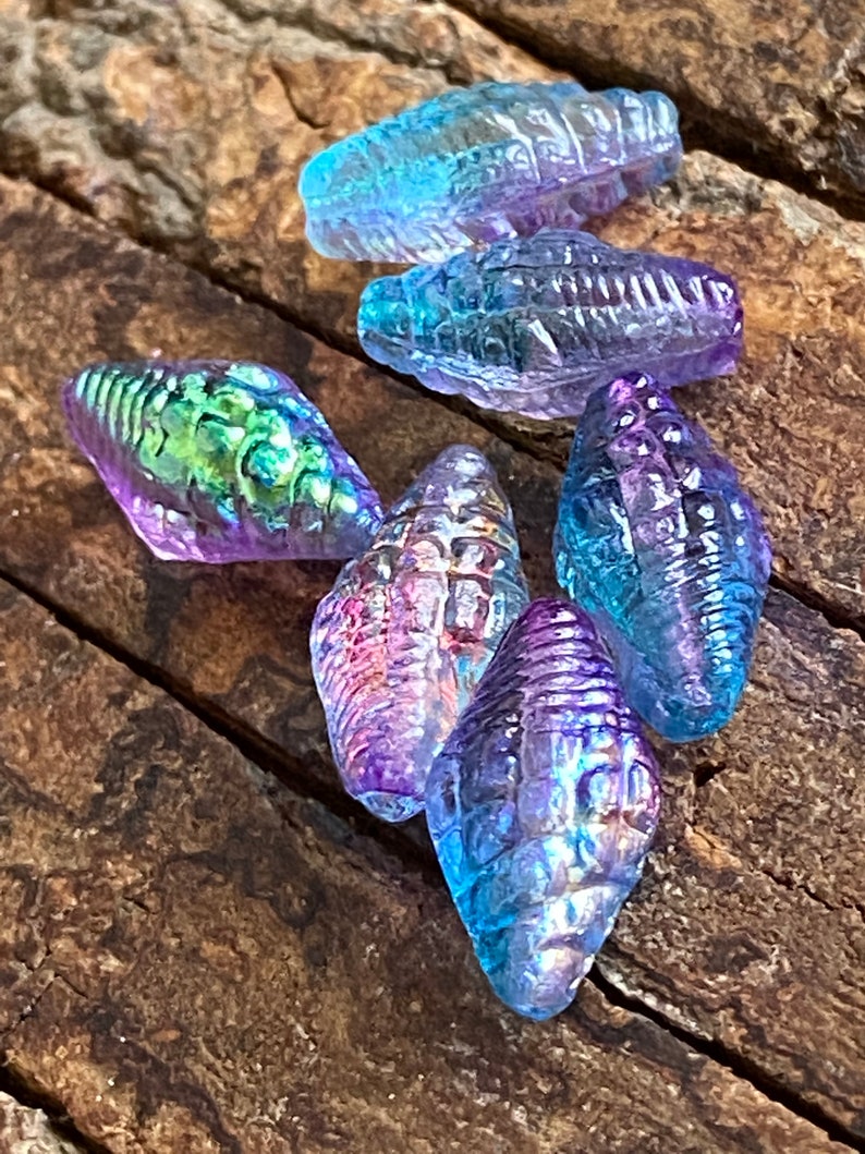 May include: A collection of iridescent glass seashell beads in shades of blue, purple, and green. The beads are all different shapes and sizes, and they have a textured surface.