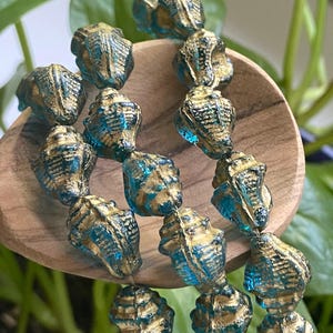 May include: A strand of seashell-shaped beads. The beads are a translucent teal color with gold accents. The beads are strung together, creating a necklace or bracelet.