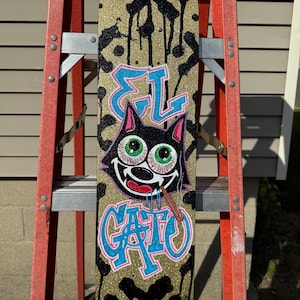 May include: A skateboard with a gold glitter background and black paint drips. The board features a cartoon cat with green eyes and the words "EL GATO" in blue and pink. A lit cigarette is in the cat's mouth.