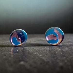 May include: A pair of circular stud earrings featuring a smooth, polished surface with a swirling pattern of translucent light blue and soft pink colors. The earrings have a rounded, dome-like shape that reflects light across their glossy finish.