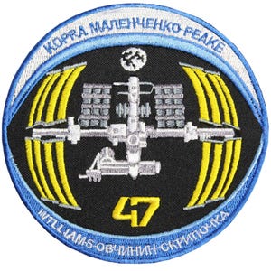 May include: A black and blue circular patch with a white outline. The patch features a white and gray illustration of the International Space Station with yellow solar panels. The text "KORRA MALENCHENKO PEAKE" is written in a white arc above the illustration. The text "WILLIAMS OBLIČININ SKRIPOČKA" is written in a white arc below the illustration. The number "47" is written in yellow in the center of the patch.