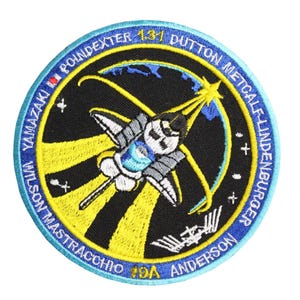 Puede incluir: Un parche bordado negro y amarillo con una lanzadera espacial que se lanza hacia una estrella. El texto alrededor del borde dice "STS-131 DUTTON METCALF LINDENBURGER ANDERSON YAMAZAKI POINDEXTER MASTRACCHIO 19A".