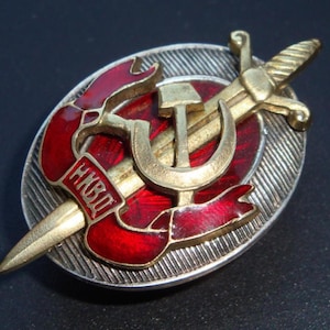 May include: A gold and red enamel pin with a crossed hammer and sickle, a sword, and a red ribbon with the text "НКВД" in Cyrillic script.