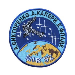 May include: A blue and yellow embroidered patch with the text "АВІКТОРЕНКО А.КАПЕРЯК КОСМОНАВТ" and "MIR 92" in the center. The patch depicts a spacewalk with a cosmonaut and the Earth.