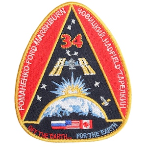 Può includere: Un cerotto triangolare blu e rosso con il testo "ROMANENKO-FORD-MARSHBURN" e "CHOVIKIN-HADFIELD-TAPEJKIN" sui lati. Il centro del cerotto presenta un'immagine stilizzata della Terra con una stazione spaziale in orbita sopra di essa. Il testo "OFF THE EARTH... FOR THE EARTH" è nella parte inferiore del cerotto.