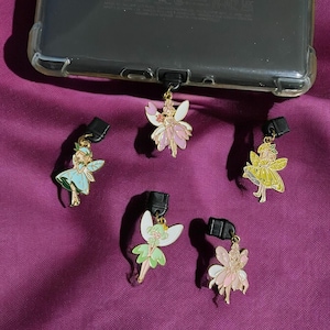 May include: Five gold-colored fairy charms with colorful wings and dresses. The charms are attached to black phone case connectors.