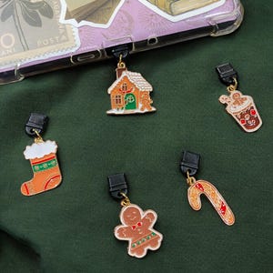 May include: Five gold-colored Christmas-themed charms with black clips. The charms include a gingerbread house, a gingerbread man, a candy cane, a stocking, and a cup of hot chocolate.
