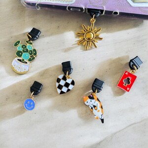 May include: A set of six phone charms featuring a variety of designs, including a sun, a potted plant, a heart, a cat, a smiley face, and a playing card. The charms are made of metal and have a gold finish.
