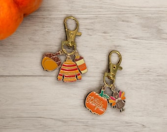May include: Two gold-toned metal keychains featuring autumn-themed charms. One keychain has an acorn, a striped sweater, and a coffee cup. The other keychain features an orange pumpkin with the text Hello Pumpkin and a colorful turkey charm.