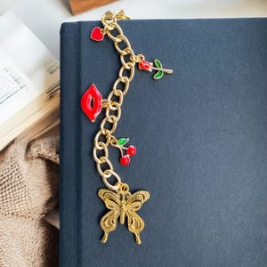 May include: A gold chain bookmark with red and green enamel charms, including a heart, a rose, lips, a cherry, and a butterfly.