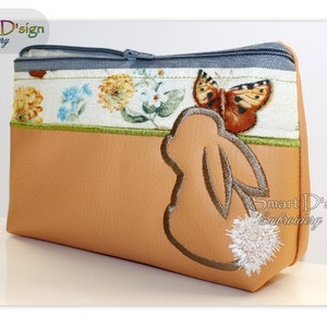 May include: A tan zippered cosmetic bag with a floral pattern and a butterfly applique. The bag features a bunny silhouette with a fluffy white tail. The top is trimmed with a blue zipper and a green border.