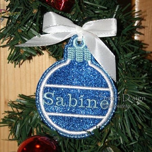 May include: A blue and white Christmas ornament with the name "Sabine" embroidered on it. The ornament is hanging from a white ribbon on a green Christmas tree.