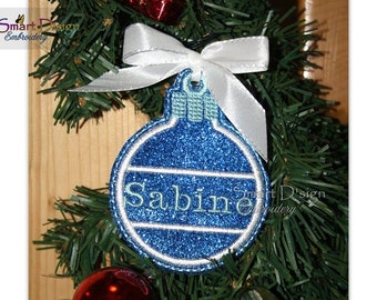Op de afbeelding: Een blauwe en witte kerstbal met de naam "Sabine" erop geborduurd. De bal hangt aan een wit lint aan een groene kerstboom.