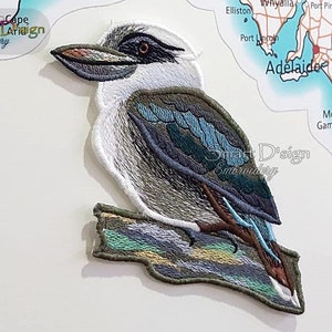 May include: Embroidered patch of a kookaburra bird perched on a branch. The bird is brown, blue, and white with a long beak. The branch is green, blue, and yellow.