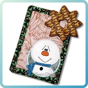 Puede incluir: Un artículo de tela rectangular con temática navideña con un aplique de muñeco de nieve y una galleta en forma de estrella. El muñeco de nieve tiene una bufanda azul y una nariz naranja. El fondo tiene un estampado floral.