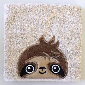 Op de afbeelding: Een beige handdoek met een geborduurd bruin luiaardgezicht. De luiaard heeft grote zwarte ogen met witte pupillen en een kleine, gesloten glimlach.