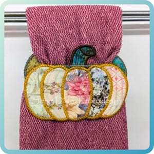 May include: A hand towel with a stitched pumpkin design. The pumpkin is made of fabric with floral patterns and a gold trim. The towel is a textured, dusty rose colour.