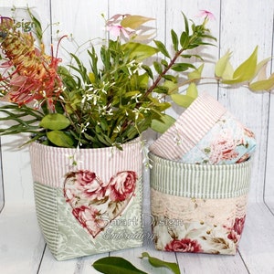 Könnte beinhalten: Zwei Stoffaufbewahrungskörbe mit Blumenmustern und rosa-grünen Streifen. Die Körbe sind mit Blumen gefüllt und haben ein herzförmiges Design auf der Vorderseite. Der Text "Smart Design Embroidery" ist auf den Körben sichtbar.