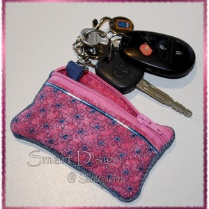 May include: A pink fabric key chain with a zipper closure and a blue trim. The key chain has a floral pattern and the text "Smart D'sign @Stickzauber" is embroidered on the front.