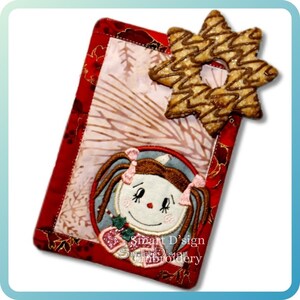 May include: A red and gold fabric bookmark featuring a doll face and a star-shaped biscuit. The bookmark has a doll face with pigtails and a Christmas holly design. The star biscuit has a chocolate drizzle.
