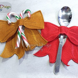 Puede incluir: Dos diseños de lazos de Navidad bordados. Uno es dorado con bastones de caramelo y el otro es rojo. El lazo rojo está envuelto alrededor de una cuchara de plata.