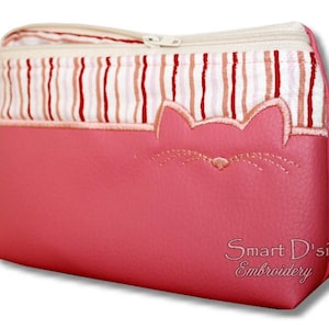 May include: A pink cosmetic bag with a cat face design. The bag features a striped pattern at the top and a zipper closure. The cat face is embroidered on the front. The text "Smart D'sign Embroidery" is visible.
