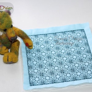 Puede incluir: Un cuadrado de tela azul con un diseño bordado en negro de formas geométricas repetitivas. El diseño es similar a un enrejado. Un pequeño animal de peluche está en primer plano.