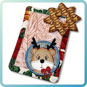 May include: A rectangular Christmas-themed quilted item with a reindeer applique and a star-shaped cookie. The reindeer has a red bow and is inside a blue circle. The item has a pink background with a leaf pattern and red and green borders.
